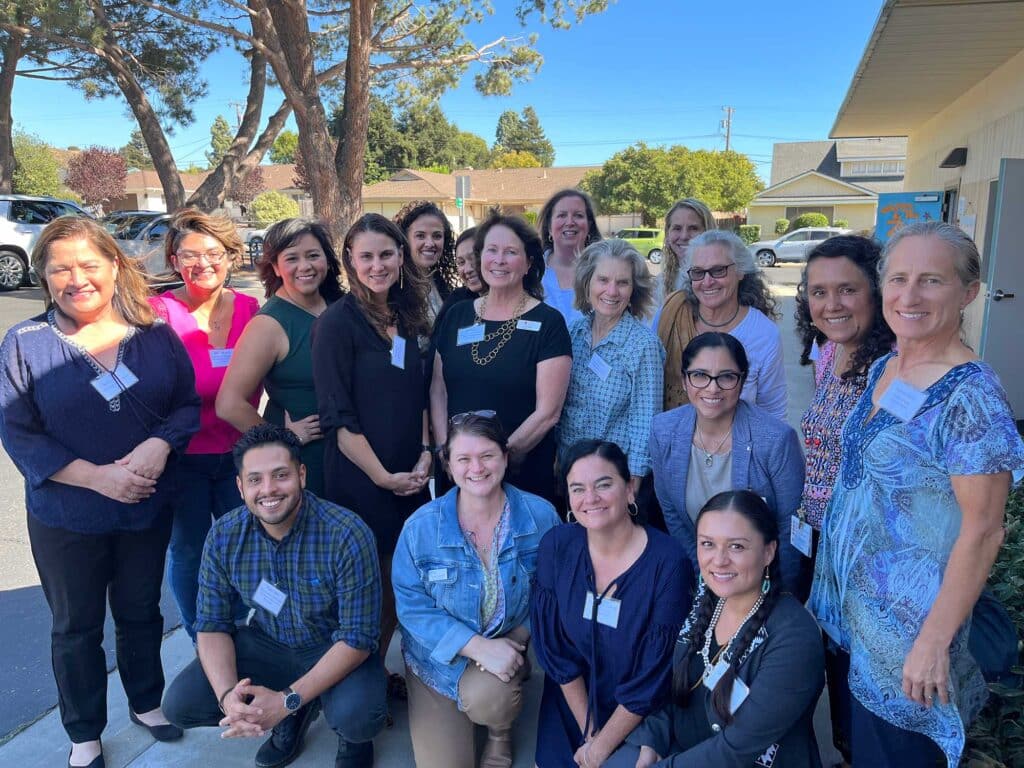 Group photo of smiling community health workers and coordinators gathered outdoors on a sunny day, standing and kneeling together in front of a building.